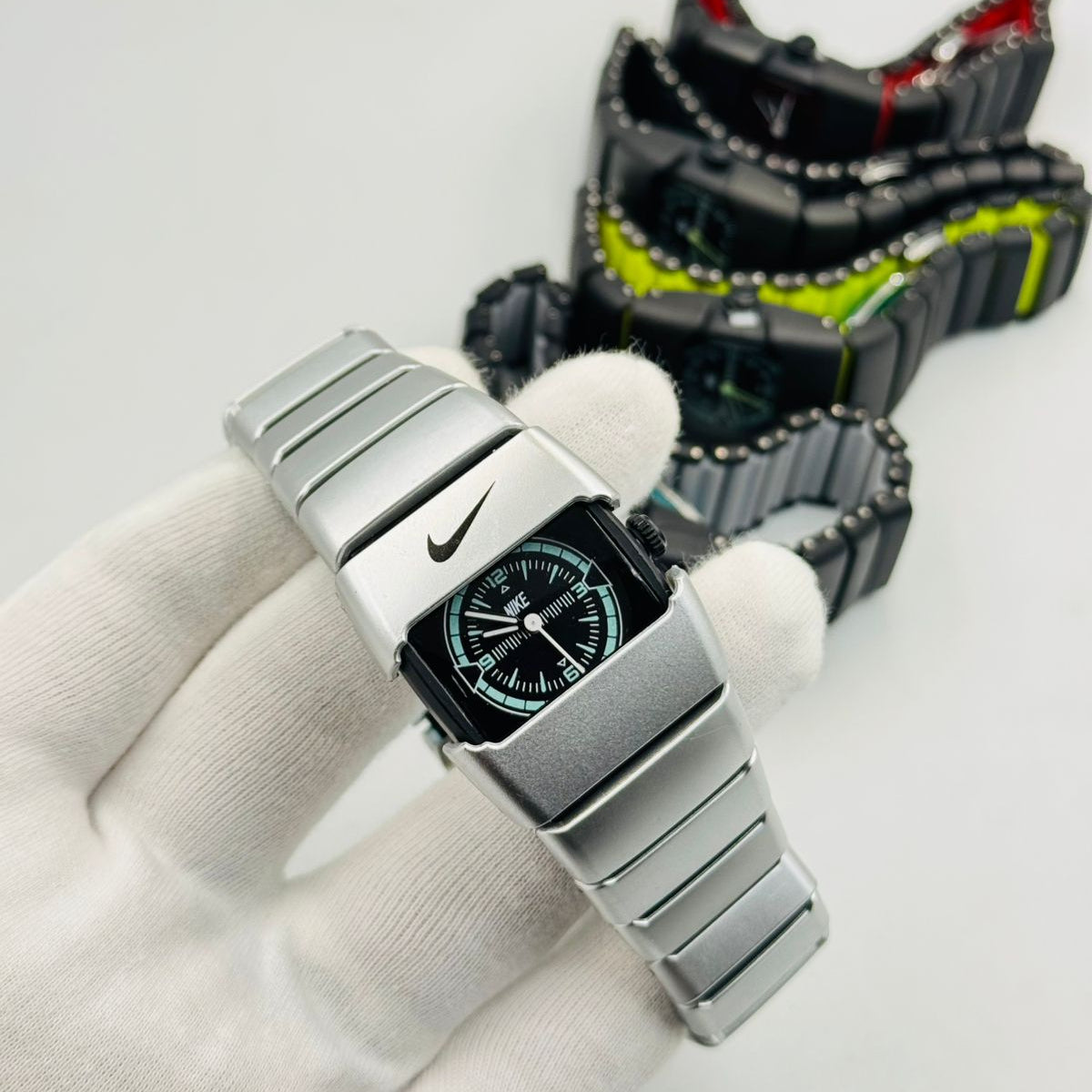 Silver watch with a black face held by a gloved hand, with another watch in the background on a white surface.