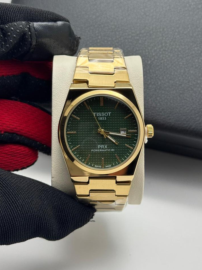 Gold wristwatch with green face on a white surface, surrounded by black gloves and a gray box.