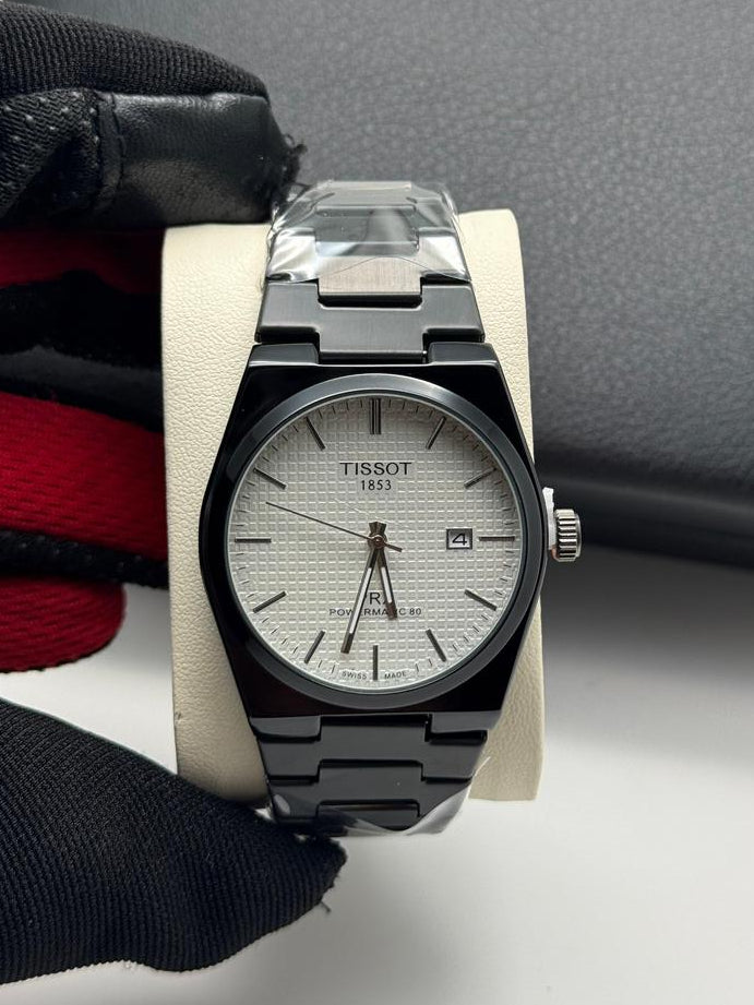 Tissot watch with black and silver band held by a gloved hand against a gray background