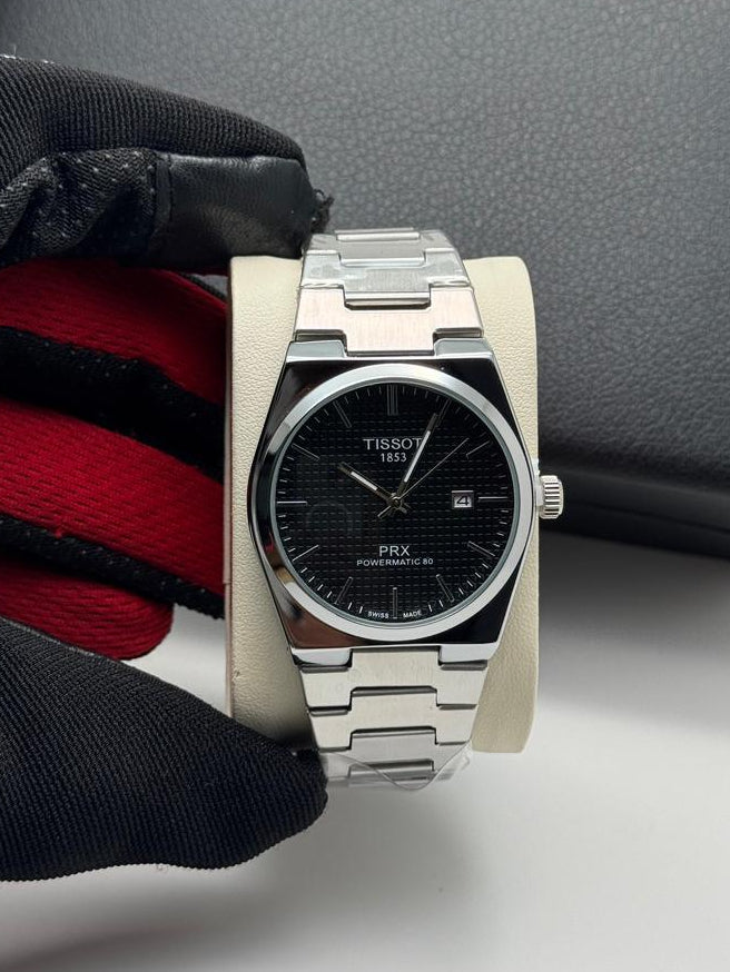 Silver Tissot watch with black face on a white surface, next to a black box.